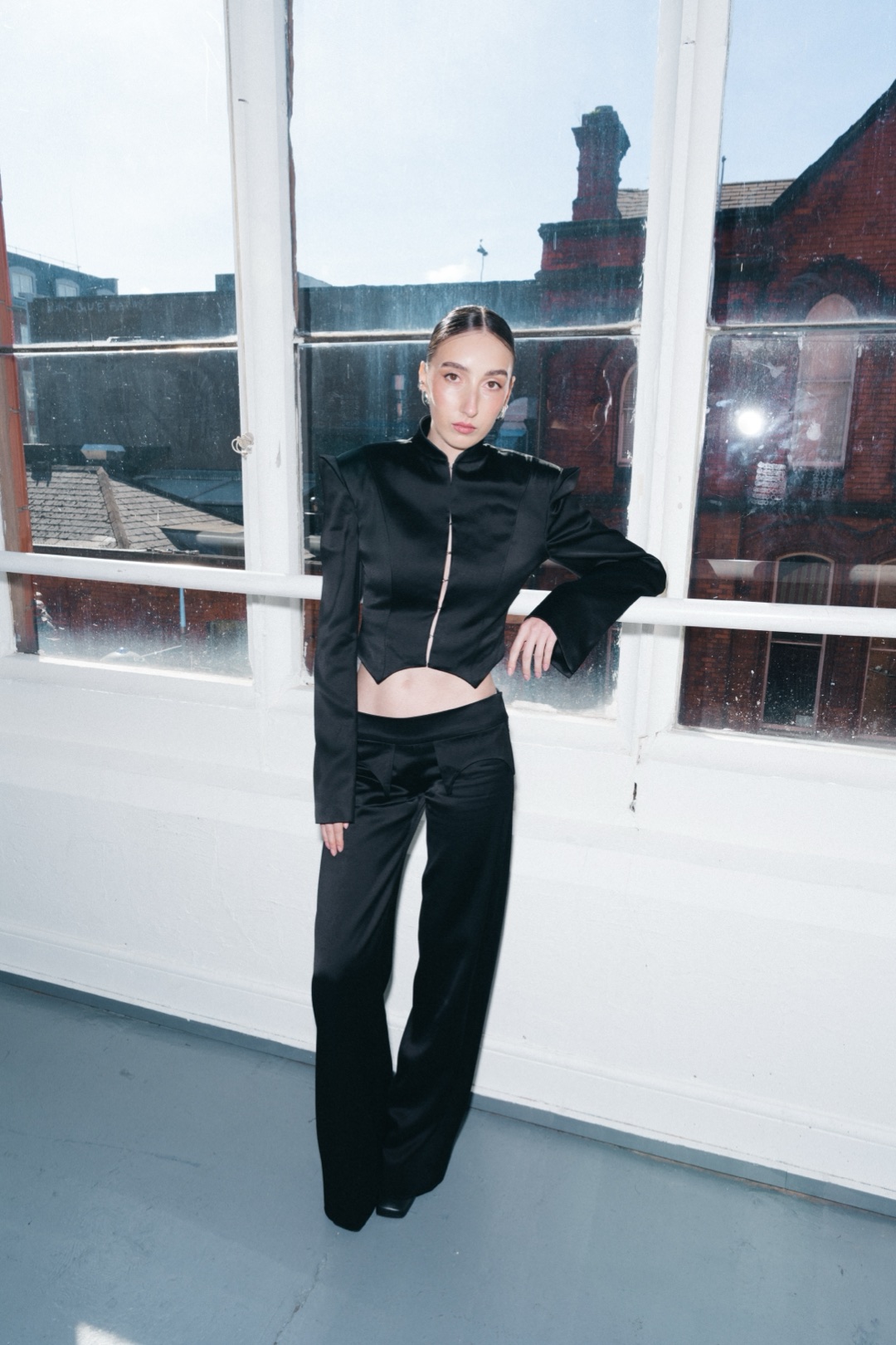 The black suit, full length, leaning on a window rail over red brick buildings