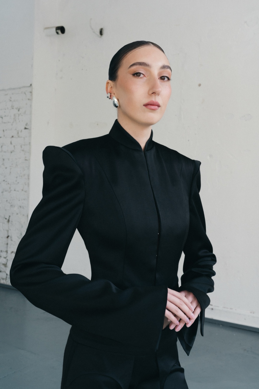 The black suit, stand collar and sculptural shoulder, hands clasped at the waist