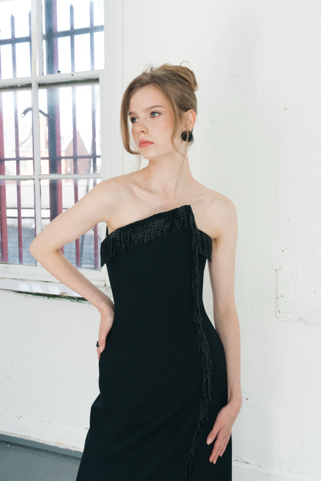 The fringe gown, waist up, hand on hip, beside a barred window